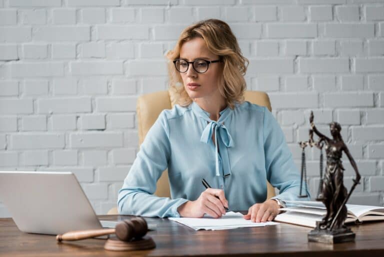 serious female judge in eyeglasses working with papers and using laptop at workplace